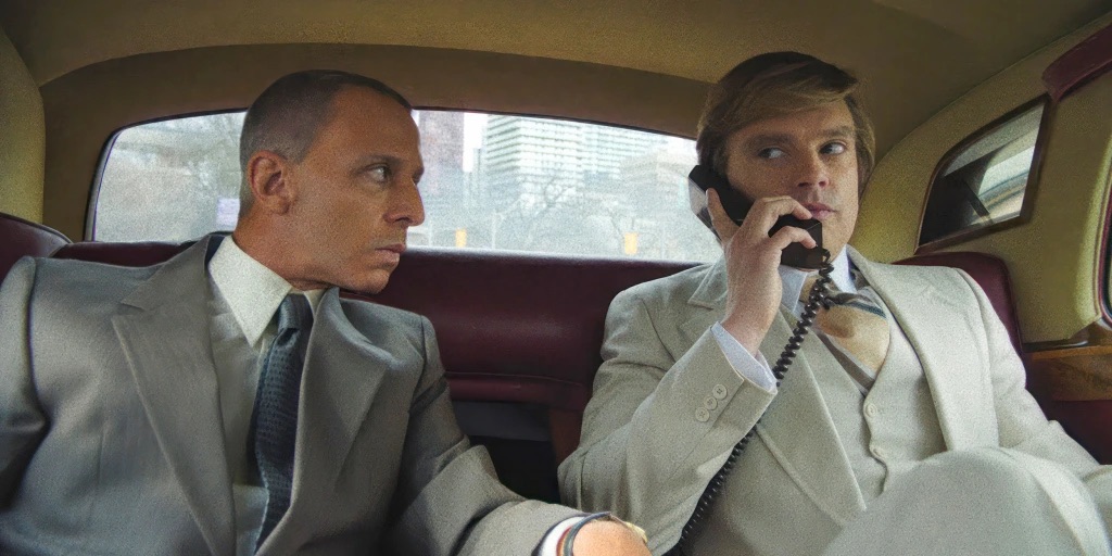 A movie still from 'The Apprentice' (2024). Roy Cohn (Jeremy Strong) and a young Donald Trump (Sebastian Stan) sit in the back of a luxury limousine. Cohn is looking intently at Trump, who is holding a car phone to his ear, dressed in a light-colored three-piece suit.
