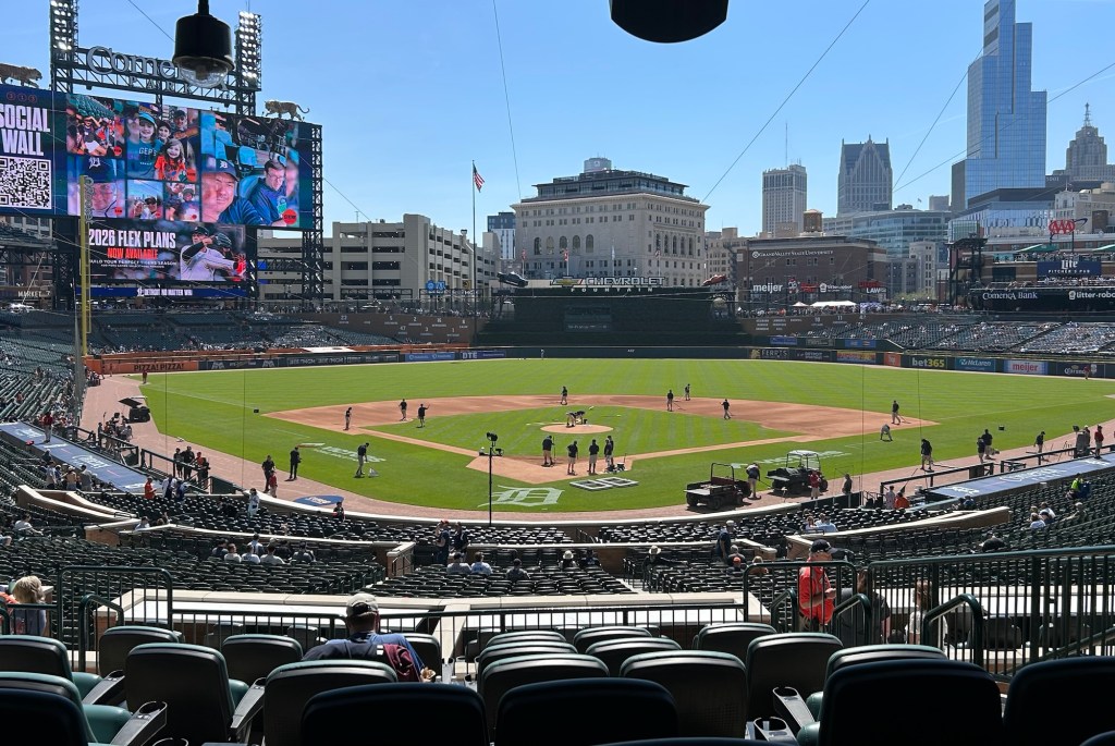 A wide-angle view of Comerica Park from the Tiger Den seats during a sunny afternoon game. The green field is perfectly manicured, the Detroit skyline is visible in the background, and the scoreboard shows fans on the social wall.