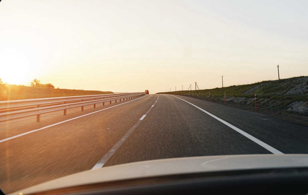 A first-person view from inside a car looking through the windshield at a long, empty two-lane asphalt highway stretching into a bright sunset on the horizo