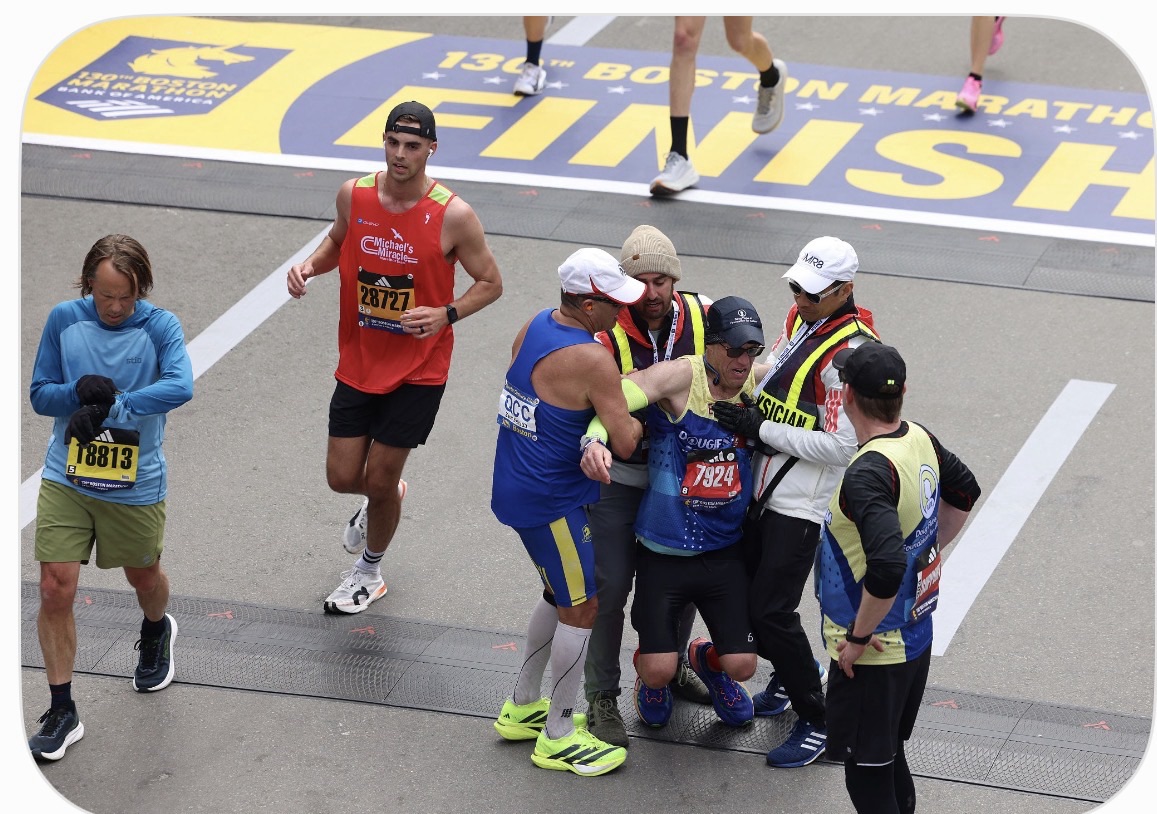 An overhead view of the Boston Marathon finish line on a grey pavement street. A runner in a blue shirt has collapsed and is being held up under his arms by race officials in high-visibility vests and a fellow runner in a blue jersey. In the background, other runners continue past the yellow and blue "Boston Marathon Finish" banner, illustrating the exact moment of choice between finishing a race and stopping to help someone up.