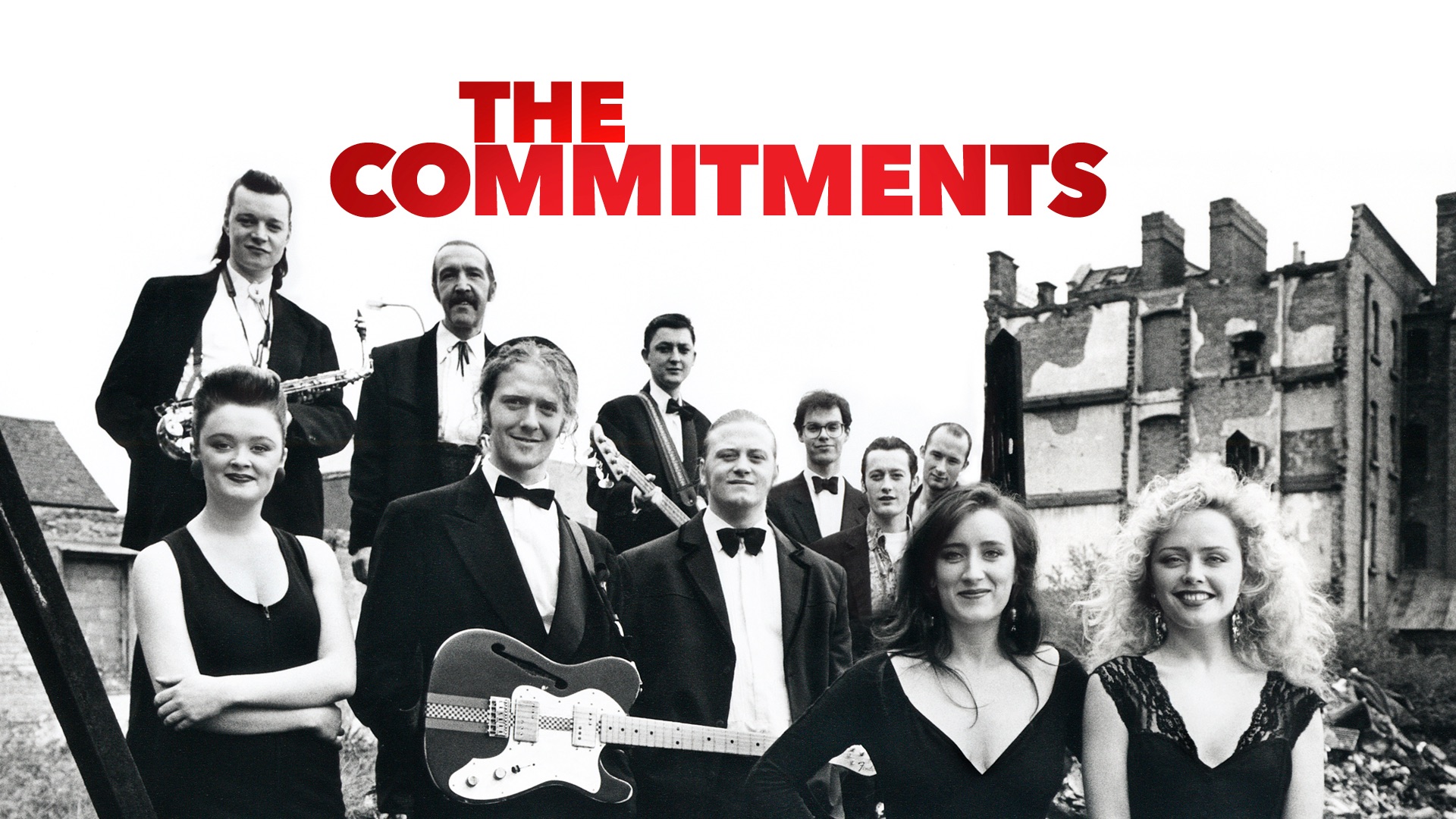 A wide, black-and-white promotional still of the cast of The Commitments in suits and formalwear outdoors against a backdrop of urban Dublin ruins, with the bold red title "THE COMMITMENTS" stamped across the top.