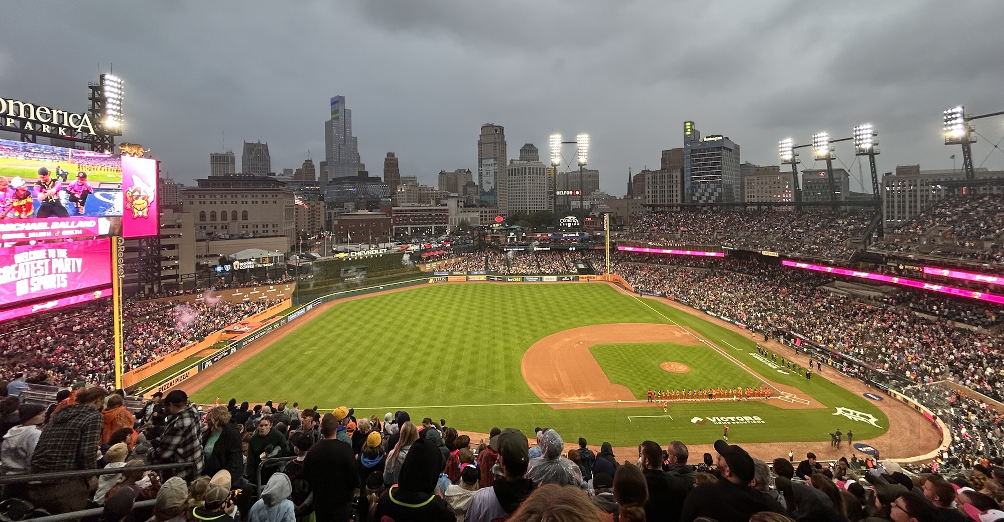 A high-angle view of a packed Comerica Park under a heavy, overcast evening sky, highlighting the bright green infield turf, vibrant stadium lights, and the downtown Detroit skyline rising in the background.