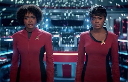 Medium shot of actor Celia Rose Gooding playing two versions of Nyota Uhura standing on a starship bridge with a blurred, illuminated background, wearing maroon and black Starfleet uniforms with gold command insignia pins.