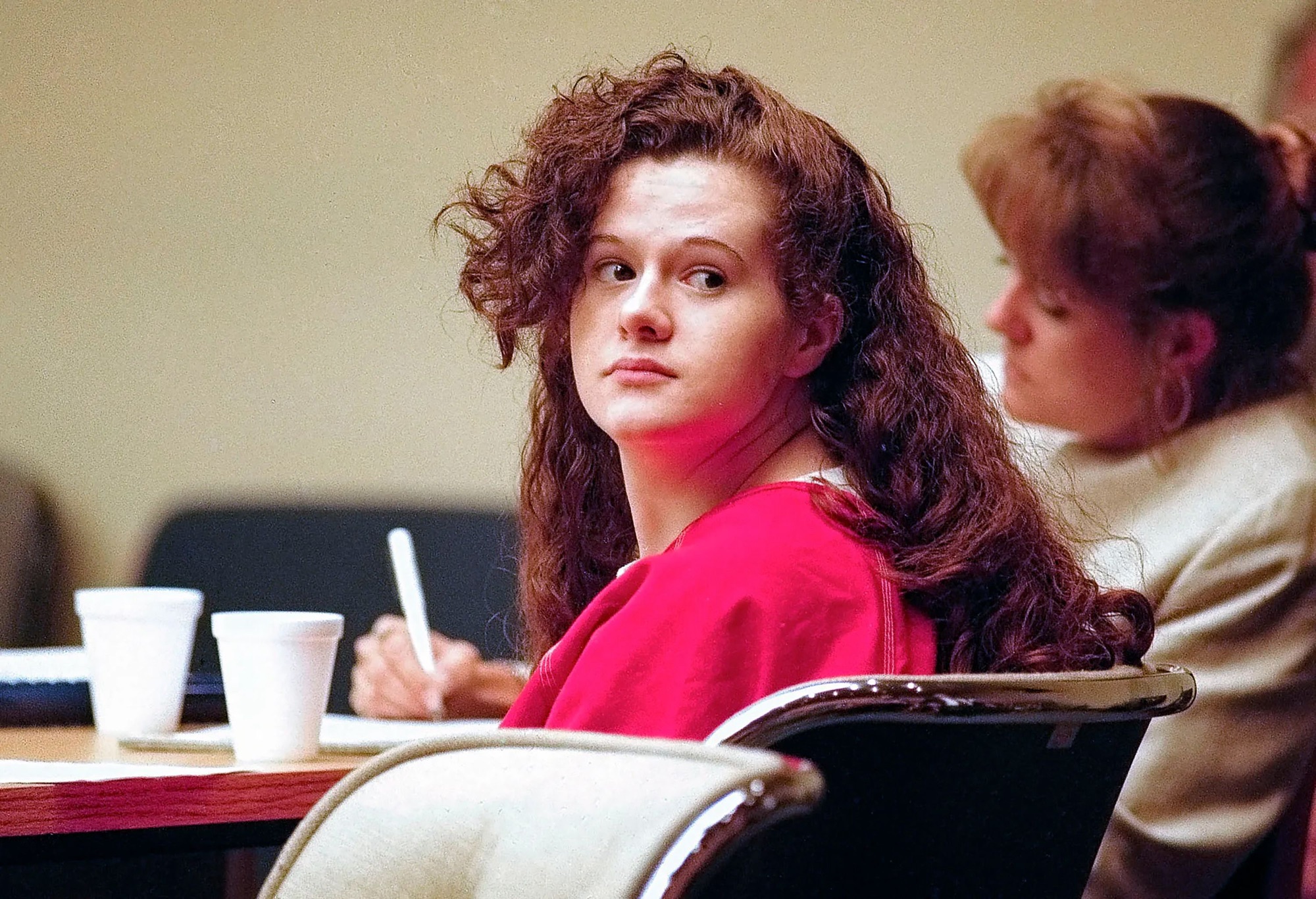 Archival courtroom photograph of Christa Pike with long curly reddish-brown hair, wearing a red jail uniform and looking back over her shoulder while writing at a table during her 1996 trial.