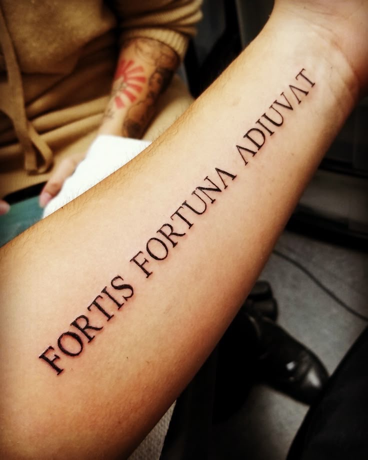A close-up photograph of a clean, black-ink forearm tattoo in a serif font. The Latin phrase "FORTIS FORTUNA ADIUVAT" (Fortune favors the bold) is clearly visible and sharply in focus. The arm rests naturally, serving as a physical testament to the life philosophy discussed in the post.
